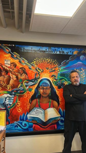 Joseph Nuke Montalvo posing in front of his mural, Flor y Canto, in the Chicanx/Latinx Student Center.